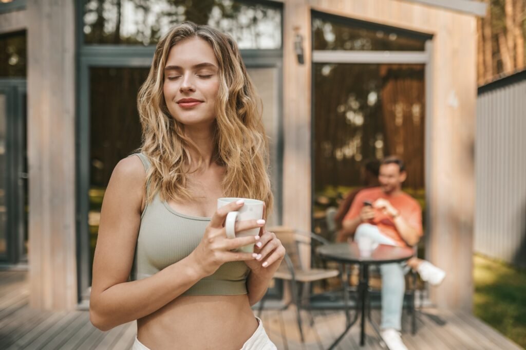 Pretty long-haired blonde woman with a coffee cup standing outsie the cottage