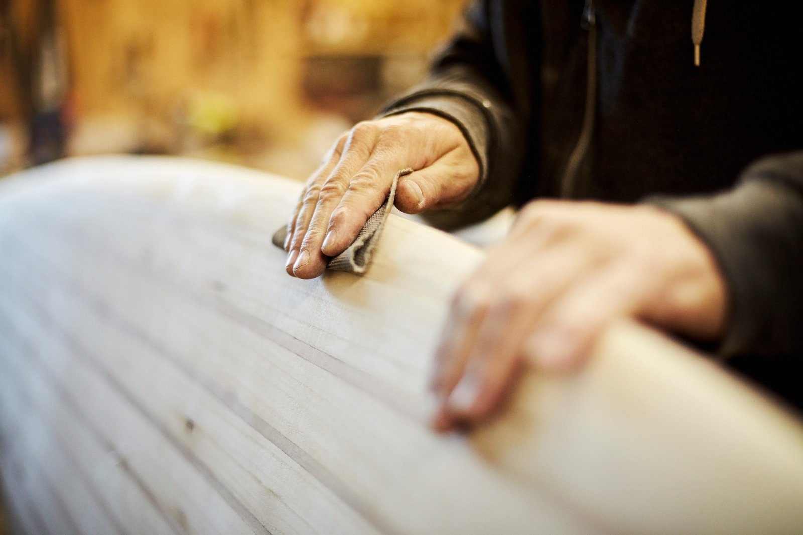 Man standing in a workshop sanding the edge of a wooden surfboard.